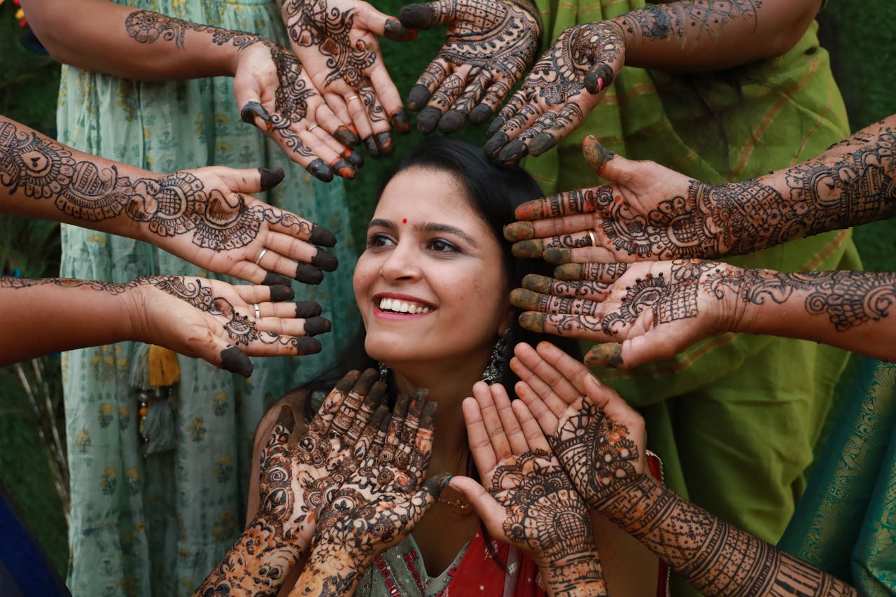 Indian bride smiling surrounded by hands with intricate henna designs, signifying cultural celebration.